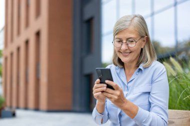 Gülümseyen, gözlüklü olgun bir kadın modern bir binanın önünde otururken akıllı telefonuna bakar. Mutlu ve rahat görünüyor..
