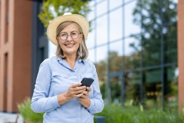 Şapka takan zarif, yaşlı bir kadın telefonunu açık havada tutarken gülümsüyor ve memnuniyetini gösteriyor..