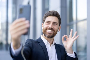 Takım elbiseli gülümseyen bir iş adamı dışarıda kulaklık takarak selfie çeker ve güzel bir jest yapar. Görüntü bir pozitiflik anını yakalar..