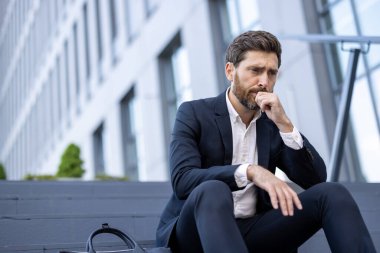 Takım elbiseli dalgın bir işadamı derin düşüncelere dalmış, dışarıda oturuyor. Yüzündeki ifade, kurumsal bir ortamda stres ya da endişe olduğunu gösteriyor. Yakınlarda bir evrak çantası var..