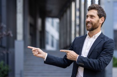Koyu takım elbiseli, gülümseyen bir işadamı iki parmağını da sağa, bir ofis binasının yakınını gösterir..