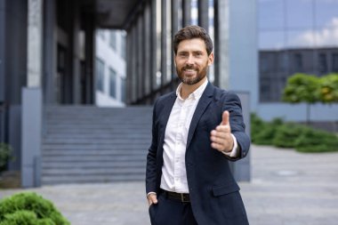 Gülümseyen bir işadamı, şık bir takım elbise giymiş ve el sıkışmaya hazır bir şekilde elini uzatır. Bu resim açık havada, iş ortamında çekilmiştir..