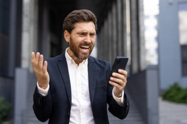 Sinirli bir işadamı telefonuna bakıp hayal kırıklığını ifade ediyor. Modern bir ofis binasının önünde duruyor..