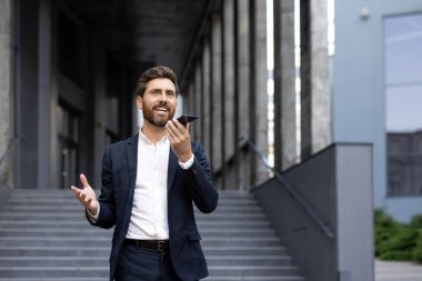 İyi giyimli bir işadamı bir şehir binasının yakınında telefonla konuşurken gülümser. Elleri sanki bir noktayı vurguluyormuş gibi hareket ediyor..