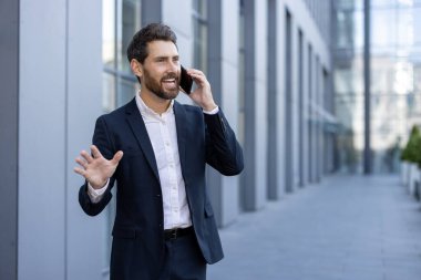 Gülümseyen bir işadamı, modern bir ofis binasının yakınında, açık havada telefonla konuşurken heyecanla el kol hareketi yapar. Keyfi yerinde gibi görünüyor..