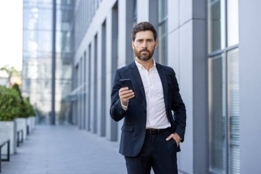 İyi giyimli bir iş adamı, akıllı bir telefonla ofis arkaplanına karşı güvenle ayakta duruyor. İletişim için hazır..