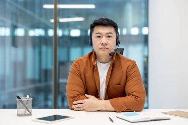 Masasında oturan Asyalı bir işadamı kameraya ciddi bir ifadeyle bakıyor. Arka plan modern bir ofis ortamını gösteriyor.