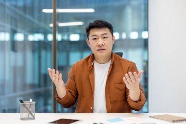 Asyalı bir işadamı konuşurken, parlak bir ofis ortamında yakalanır. O günlük turuncu bir gömlek giyiyor..