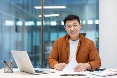 Asyalı bir işadamı masasındaki belgeler üzerinde çalışırken kameraya gülümsüyor. Modern bir ofis ortamında, iyi aydınlatılmış..