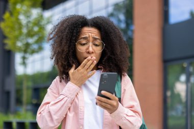 Gözlüklü genç bir kadın telefonundaki bir şeye şaşırır, elini açık havada ağzıyla kapatır. Arka planda modern bina.