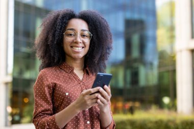 Gözlüklü genç bir kadın telefonuna bakarken gülümsüyor. Bulanık bir şehir arka planına bakıyor..