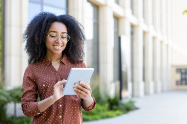 Modern bir binanın yanında tablet kullanan gözlüklü genç bir kadın gülümsüyor. Şık desenli bir gömlek giyiyor..