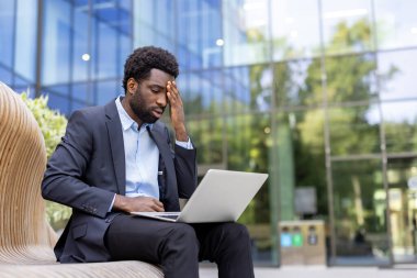 Endişeli bir iş adamı dışarıda dizüstü bilgisayarı üzerinde çalışırken başını tutuyor. Görüntü stresi ve çalışmanın zorluklarını iletir.