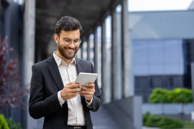 Takım elbiseli gülümseyen bir işadamı, güneşli bir günde modern bir ofis binasının önünde dikilirken tabletinde çalışır..