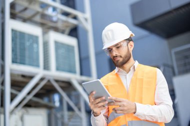 Güvenlik donanımı takan bir mühendis dışarıdaki bir tablet cihazın verilerini inceliyor, ekipmanları izliyor. Havalandırma ünitelerinin yakınındaki işine odaklanmış görünüyor..