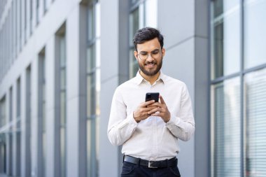 Gözlüklü, gülümseyen bir adam modern bir binanın dışında telefonuna bakıyor, mesajlaşırken mutlu görünüyor. Güneşli bir gün hoş bir manzara katar.