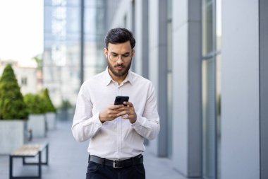 Akıllı giyimli bir işadamı, gün içinde modern bir ofis binasının önünde dolaşırken akıllı telefonunu kullanır. Kendini kaptırmış..