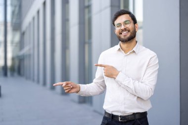 Gözlüklü, gülümseyen bir adam dışarıda durup bir şeyi işaret ediyor. Beyaz bir gömlek giyiyor ve doğrudan izleyiciye bakıyor. Arka planda modern bir bina var..