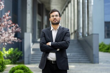 İyi giyimli bir adam güvenle dışarıda duruyor, kollarını kavuşturup, kameraya ciddi bir ifadeyle bakıyor. Arkaplanda modern bir bina yer alıyor..