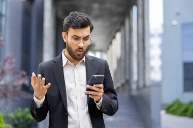 Takım elbiseli bir işadamı açık havada telefonuna bakarken şok olmuş gibi görünüyor. Telefonu elinde tutuyor ve bir binanın önünde şaşkınlıkla tepki veriyor..