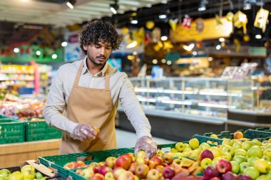 Önlük ve eldiven takan bir market çalışanı taze ürünlerden elmaları dikkatlice seçerek kalitesini kontrol eder..
