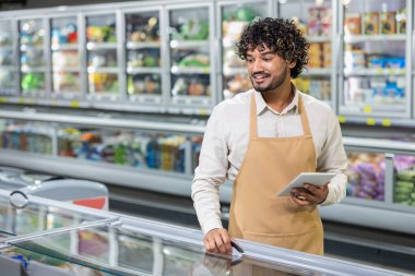 Bir market çalışanı, marketteki soğutulmuş ürünleri kontrol ederken tablet kullanarak gülümsüyor. Önlük giyiyor..