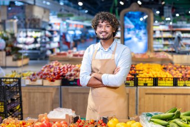 Dost canlısı bir süpermarket çalışanı taze ürünlerin önünde dururken gururla gülümser. Açık mavi bir gömlek ve bej bir önlük giyiyor..