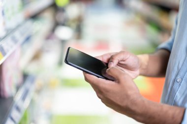 Süpermarket koridorunda akıllı telefon tutan biri, muhtemelen alışveriş yapıyor ya da fiyatları karşılaştırıyor. Yumuşak odak, teknolojiyi ve ortamı vurgular..