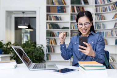 Kütüphanede heyecanlı bir kadın, etrafı kitaplarla ve teknolojiyle çevrili telefonuna bakarken bunu kutluyor..
