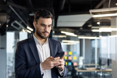 Takım elbiseli odaklanmış bir işadamı akıllı telefonunu kontrol eder, modern ofis arka planda bulanıklaşır. Mesajları ya da e-postaları okurken derin düşüncelere dalıyor..