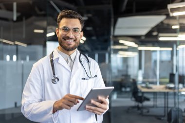 Beyaz önlüklü gülümseyen bir doktor elinde tablet bilgisayarla kameraya bakar. Modern ofis dekorasyonu resme profesyonel bir hava katıyor..