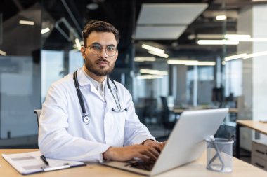 Kendine güvenen bir doktor masasında oturmuş, modern bir ofis ortamında dizüstü bilgisayar üzerinde çalışıyor. Steteskop takıyor ve kameraya bakıyor..