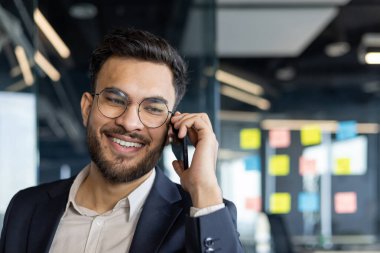 Gözlüklü gülümseyen bir adam ofis ortamında telefonuyla konuşuyor, sohbetin tadını çıkarıyor..