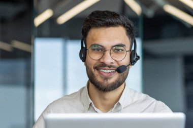 Gülümseyen bir müşteri hizmetleri temsilcisi kulaklık takıyor ve destek veriyor. Ofis ortamında dostça bir ifadeyle kameraya bakıyor..