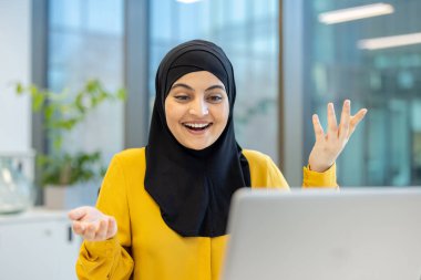 Tesettürlü neşeli bir kadın, heyecanlı bir ifadeyle, muhtemelen bir ofis ortamında dizüstü bilgisayarın önünde otururken coşkuyla el kol hareketi yapar..