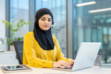Başörtüsü takan ve kulaklık takan Arap bir kadın masasındaki dizüstü bilgisayarda çalışıyormuş. Müşteri hizmetleri merkezinde çalışıyormuş gibi görünüyor..