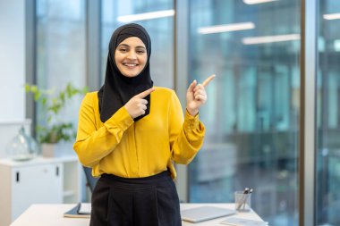 Başörtüsü içinde gülümseyen bir kadın solundaki bir şeye işaret eder. Önünde bir masa olan bir ofiste durur..