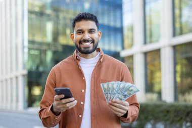 Dışarıda gülümseyen bir adam elinde nakit para ve akıllı telefon tutuyor. Açık havada başarı ya da finansal başarı öneriyor. Bina modern bir kentsel bağlam ekliyor.