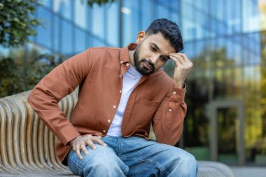 Dalgın bir adam, eli alnında, derin düşünceler içinde bir bankta oturur. Modern mimari, doğaya keskin bir zıtlık ekler..