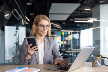Modern ofis ortamında, gözlüklü bir iş kadını telefonunu tutarken dizüstü bilgisayarıyla çalışır. Çoklu görev gibi görünüyor..