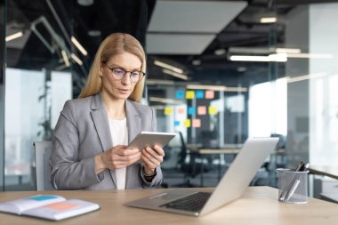 Gözlüklü bir iş kadını modern bir ofiste masa başında çalışırken bir tablet kullanır. Bir dizüstü bilgisayar ve defter de mevcut.