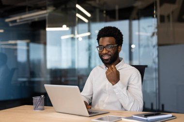 Gözlüklü, gülümseyen siyah bir işadamı elinde dizüstü bilgisayarla masa başında işe hazır bir şekilde oturuyor. Arka planda camdan duvarları olan modern ofis görülebilir..