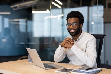 Gülümseyen siyah bir işadamı, elinde dizüstü bilgisayar, defter ve tabletle ofisinin masasında oturuyor..