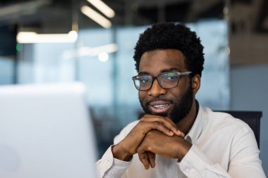 Profesyonel bir adam doğrudan kameraya bakarak sıcak bir şekilde gülümser. Gözlük takıyor ve masasında çalışmaya hazır..