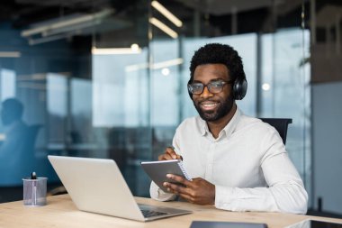 Gülümseyen bir adam kulaklık ve gözlük takıyor, ofisinde çalışıyor, kameraya bakıyor..