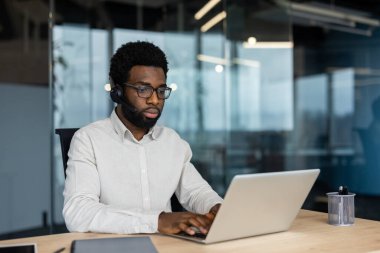 Afro-Amerikalı bir adam, modern ofis ortamında dizüstü bilgisayarı üzerinde çalışırken kulaklık takıyor ve görevlerine odaklanıyor..