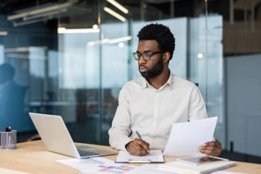 Afro-Amerikalı bir iş adamı masasında ofis ortamında bazı belgeler üzerinde çalışıyor. Yakınlarda bir laptop ve tablet var..
