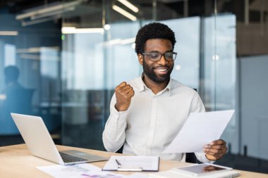 Afro-Amerikalı bir adam masasında kendinden geçmiş bir şekilde oturuyor. Elinde kağıtlar, dizüstü bilgisayarlar ve tablet var. Neşeli görünüyor ve bir başarı ifadesi var..