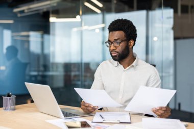 Odaklanmış bir işadamı ofis masasında bir laptop ve diğer materyalleri kullanarak belgeleri inceliyor. Profesyonel olan konsantre oluyor gibi görünüyor..