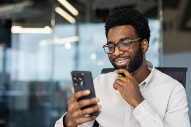 Gözlüklü yakışıklı bir adam ofis ortamında telefonuna bakarken gülümsüyor. Rahatlamış görünüyor ve anın tadını çıkarıyor..